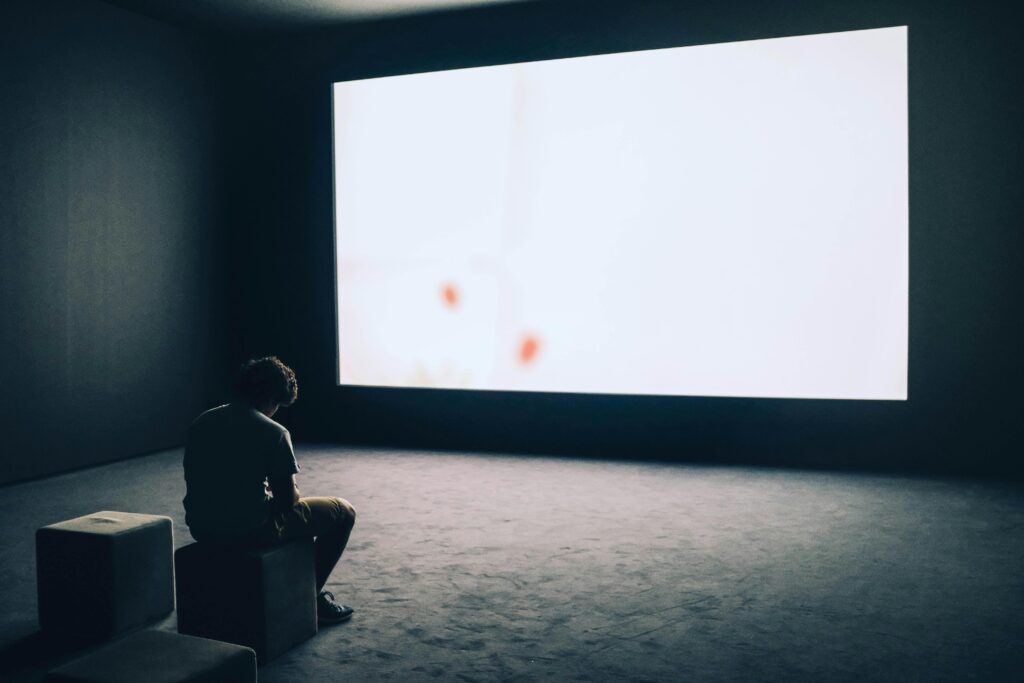 Dari Mimbar ke Layar A man sits alone in a dark room with a large white screen, depicting solitude and contemplation.